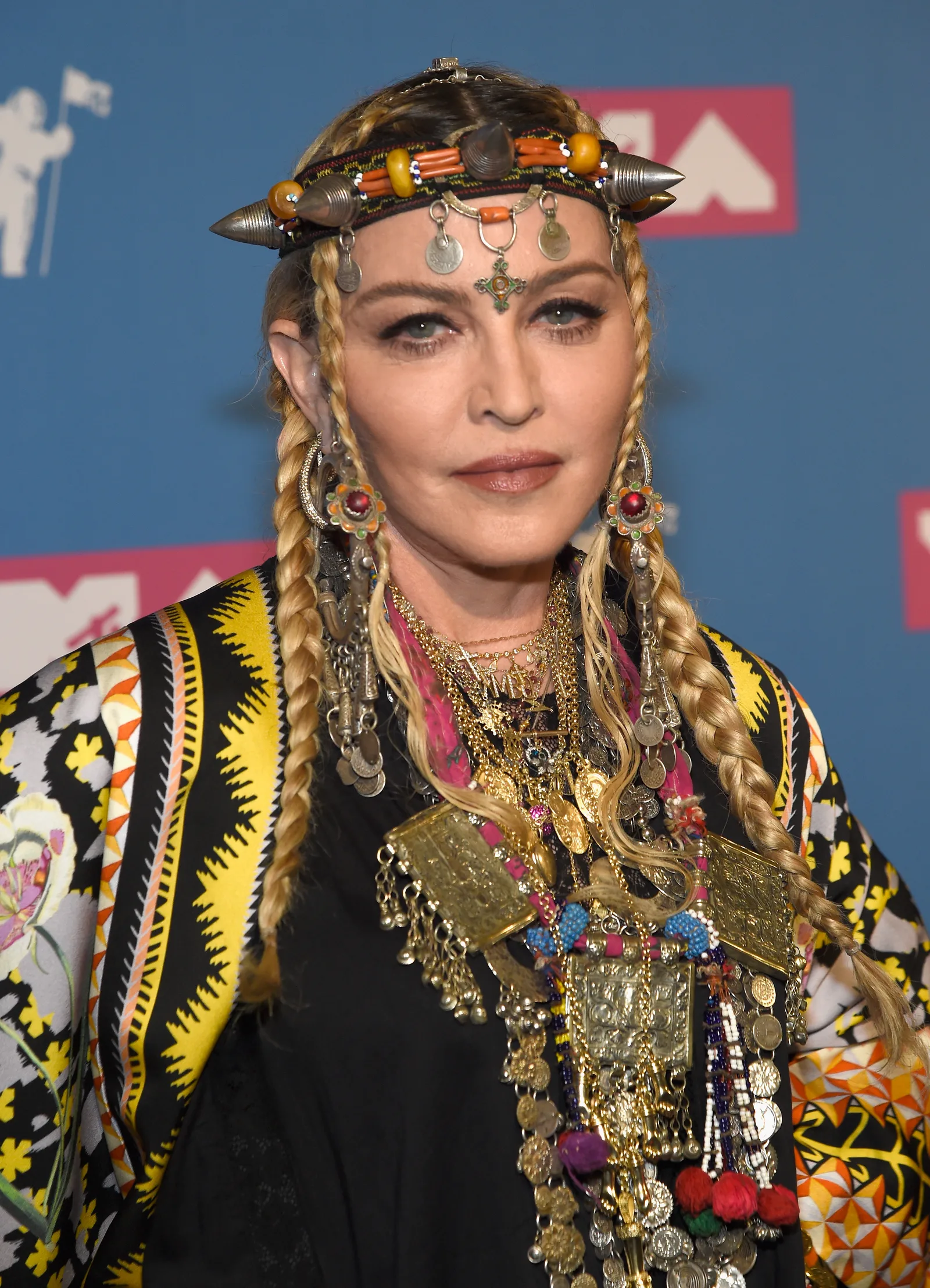 Madonna faces the camera in long blond braids, an ornate headpiece, oversized earrings, layered jewelry and a brightly patterned outfit.