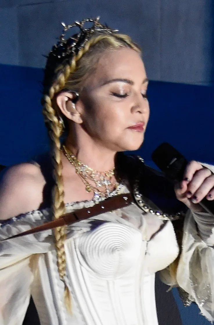 Madonna performs with long blond braids, layered necklaces and a white corset-style costume, holding a microphone as she looks downward.