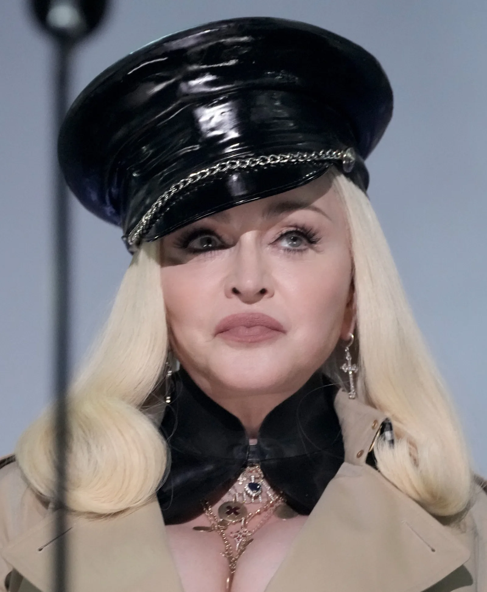 Madonna appears in a glossy black cap with chain detailing, long platinum-blond hair, dangling earrings and layered clothing and jewelry.