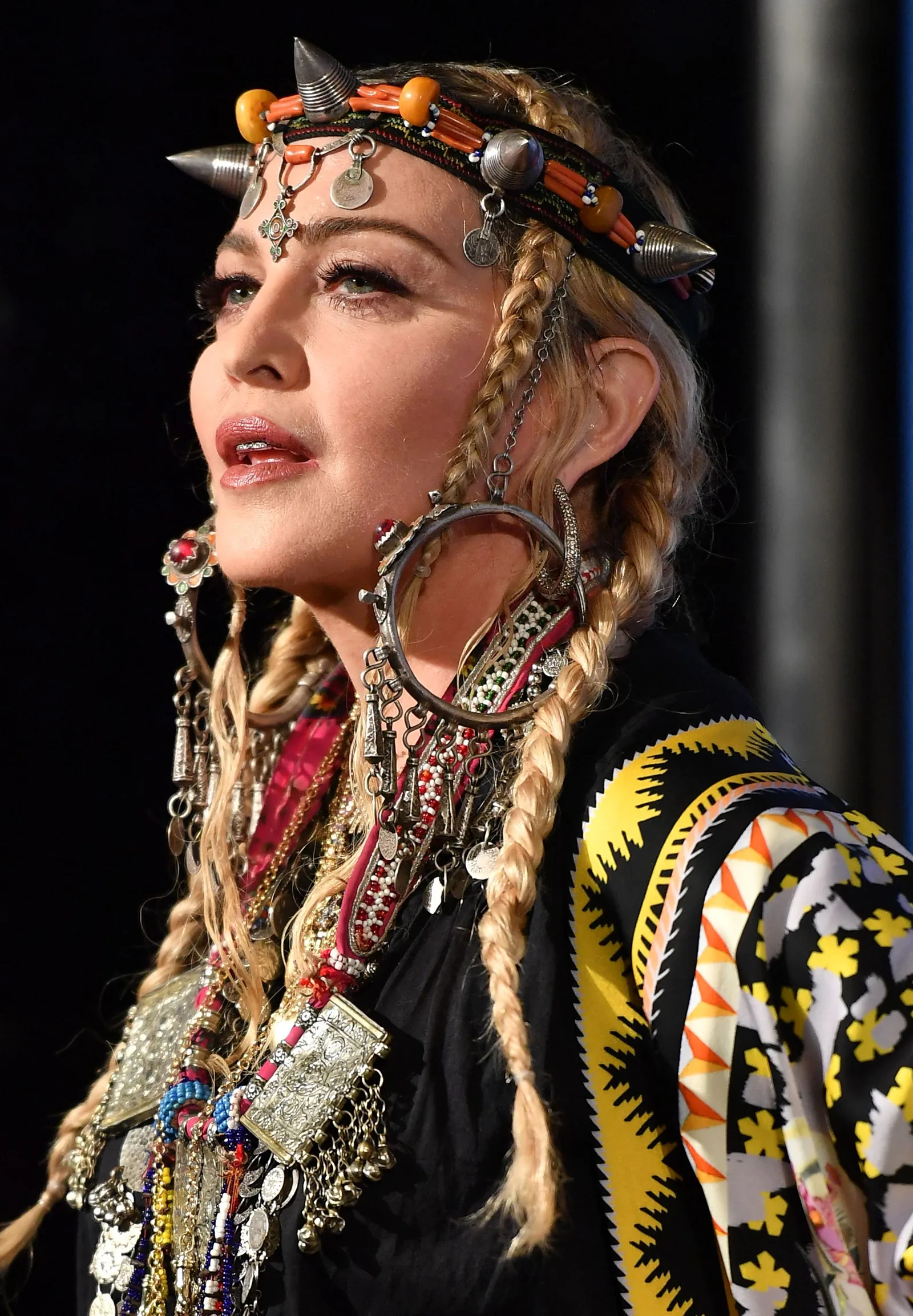 Madonna is photographed from the side wearing long blond braids, a decorative headpiece, oversized earrings and layers of intricate jewelry.