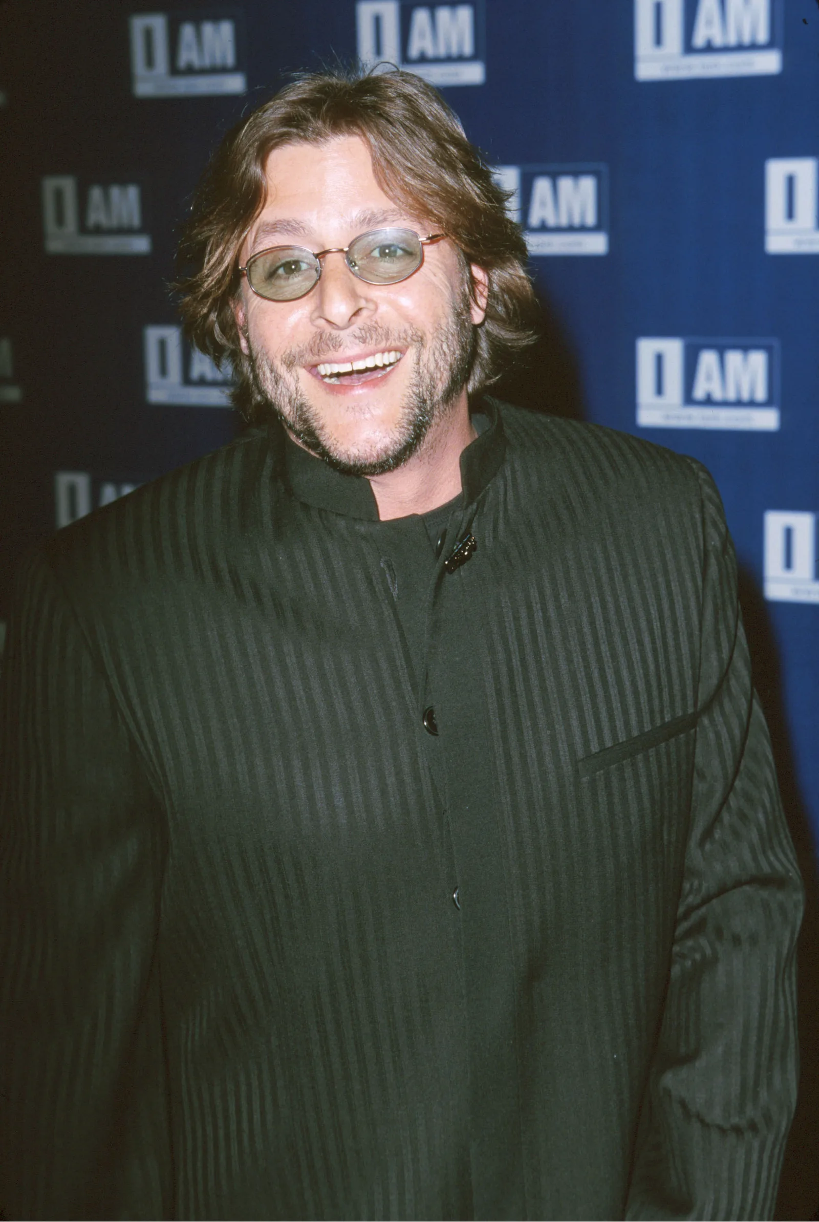 Sporting tinted oval glasses, facial stubble, and a dark pinstriped mandarin-collar jacket, the actor smiled brightly on the red carpet during the IAM.com Hollywood Launch Party at Quixote Studios in Hollywood on March 21, 2000.