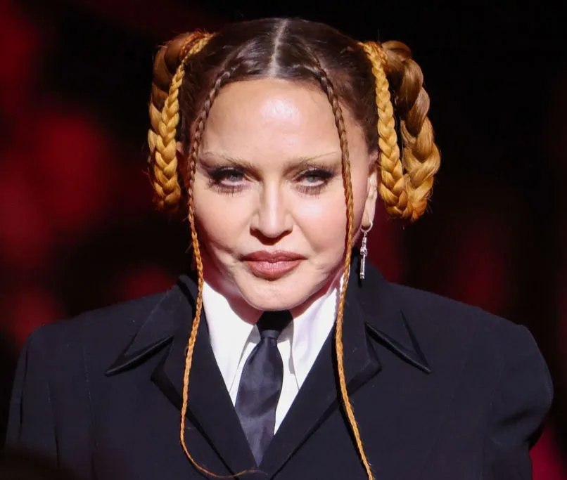 Madonna faces the camera in a black tailored suit, white shirt and black tie, with her hair styled in elaborate braided loops and long braids framing her face.