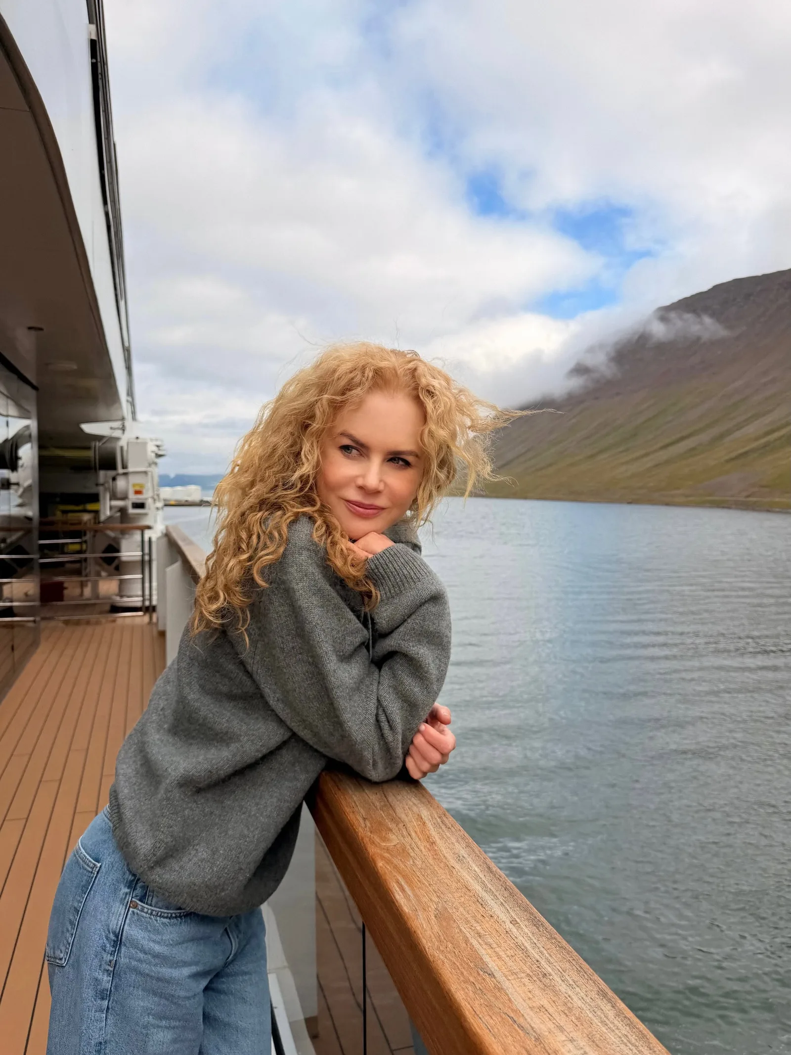 Nicole Kidman shows off her signature natural curls while leaning against the railing of a luxury Ritz-Carlton yacht off the coast of Iceland. Wearing a cozy gray sweater and denim jeans, the star embraced the windswept look during her family getaway.
