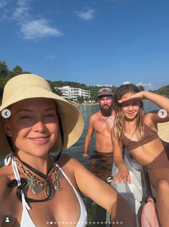 Family members gathered in the water near the shore during the European getaway, with a tree-lined coast behind them. The swimwear and seaside setting establish the warm-weather trip that later frames Goldie Hawn's appearance in the family video.