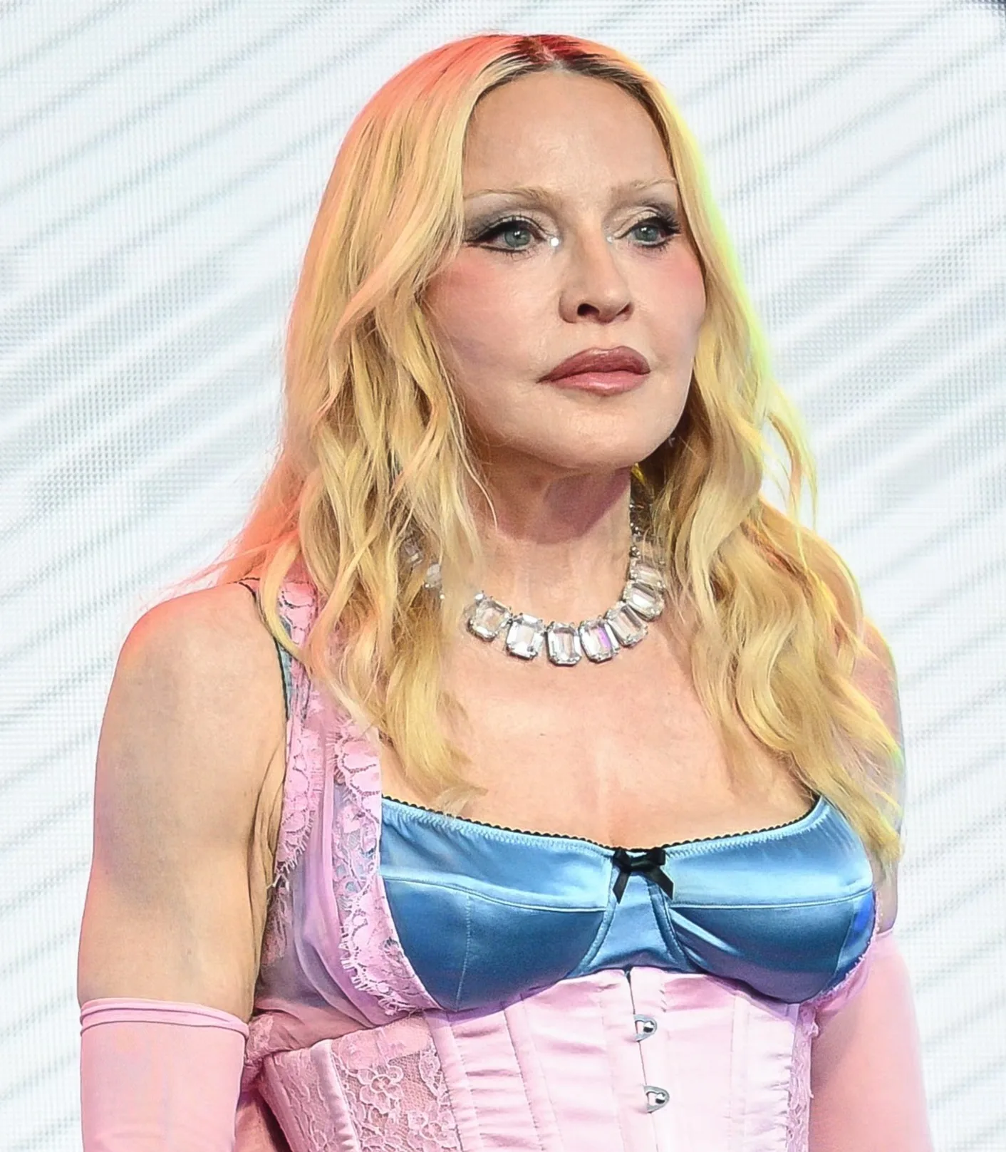 Madonna poses in a pink-and-blue corset-style outfit with long blond hair, pink gloves and a large jeweled necklace.