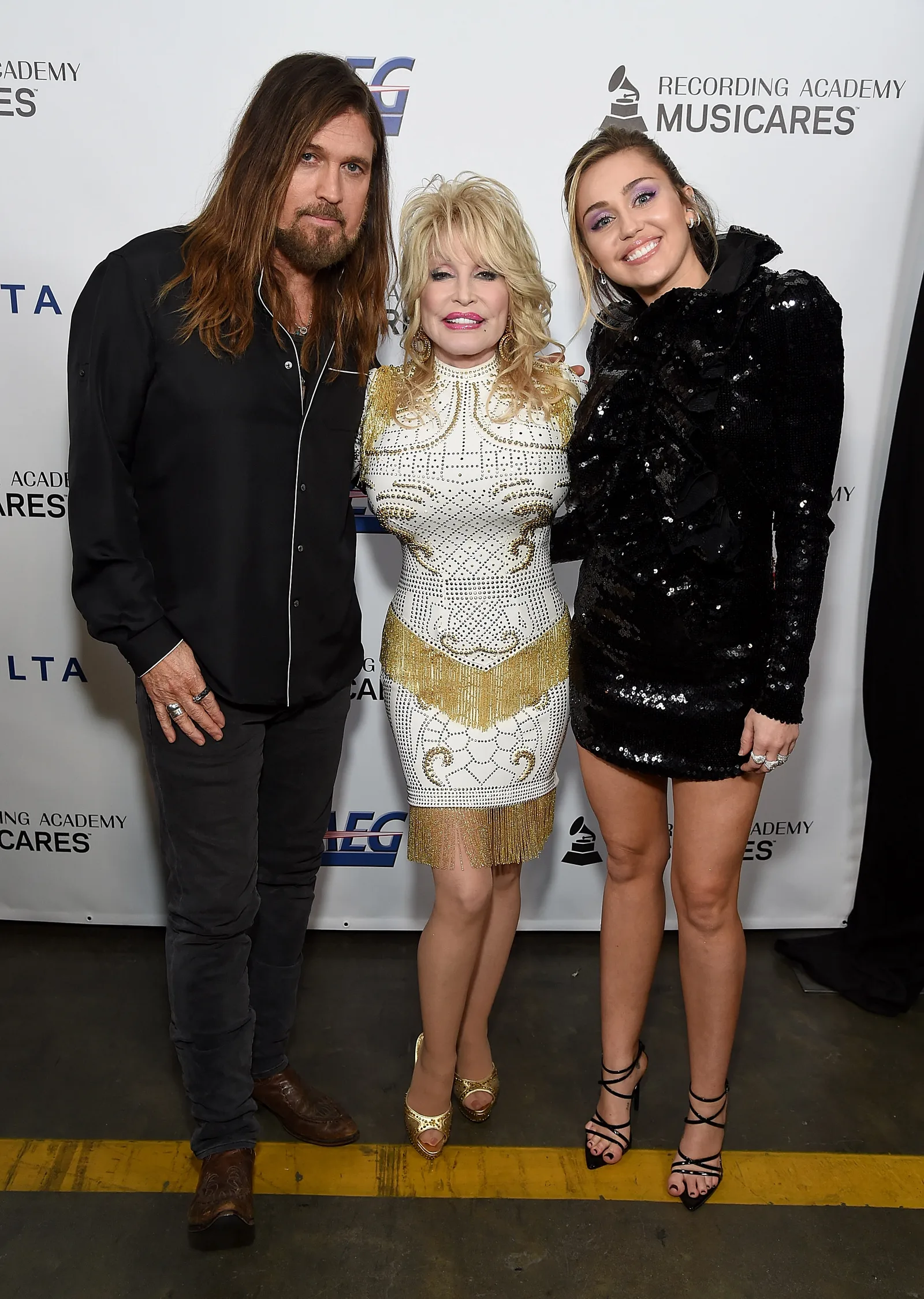 Billy Ray Cyrus, Dolly Parton, and Miley Cyrus pose together at the MusiCares Person of the Year event, with Parton standing between the father-daughter pair in a white-and-gold embellished outfit.