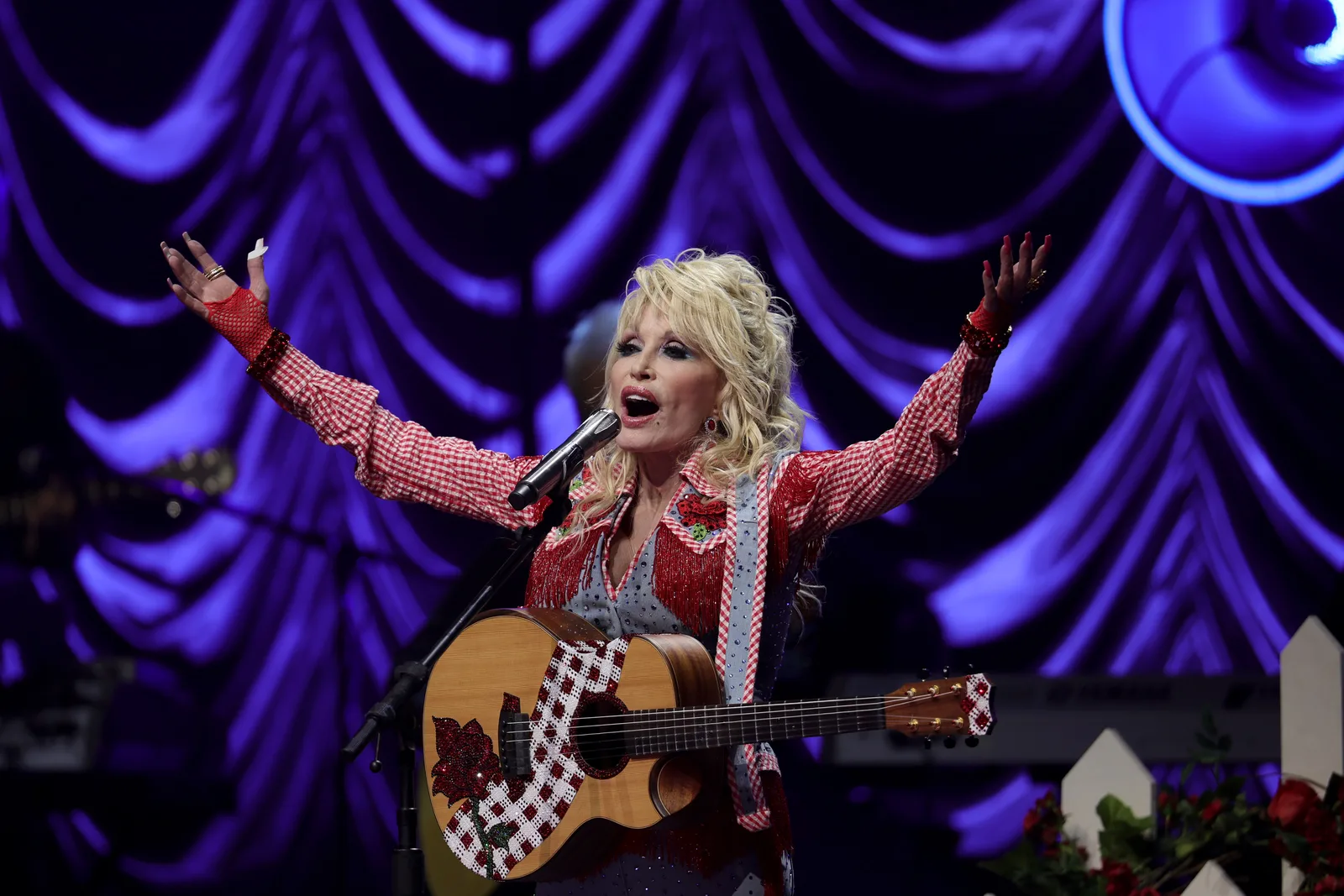 Dolly Parton performs with a guitar onstage, raising both arms as she sings into a microphone in a sparkling red-and-white outfit, with a dramatic purple-lit backdrop behind her.
