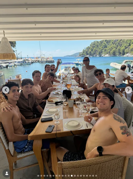 A large group sat around a long table at a waterfront restaurant. Boats and a tree-covered shoreline were visible behind them.