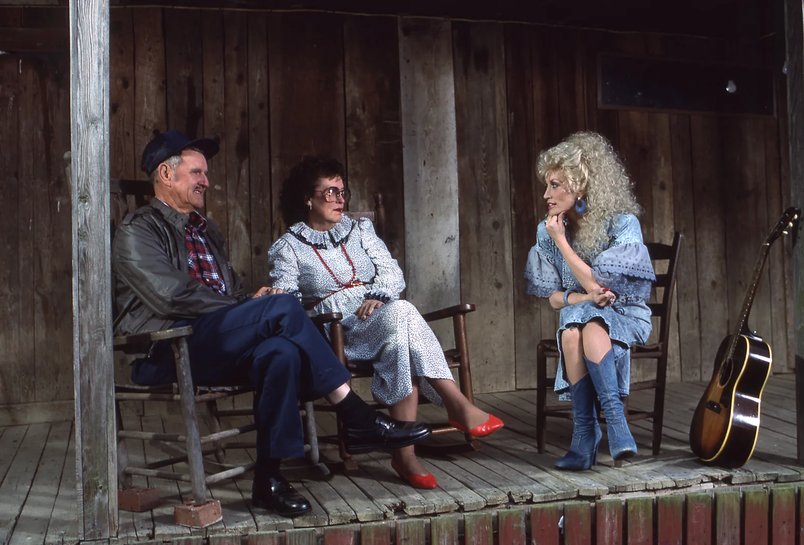 Parton is pictured with her parents, Robert Lee and Avie Lee, on the porch of the family’s rustic home, with a guitar resting nearby — a fitting glimpse of the musical roots that would shape her life.