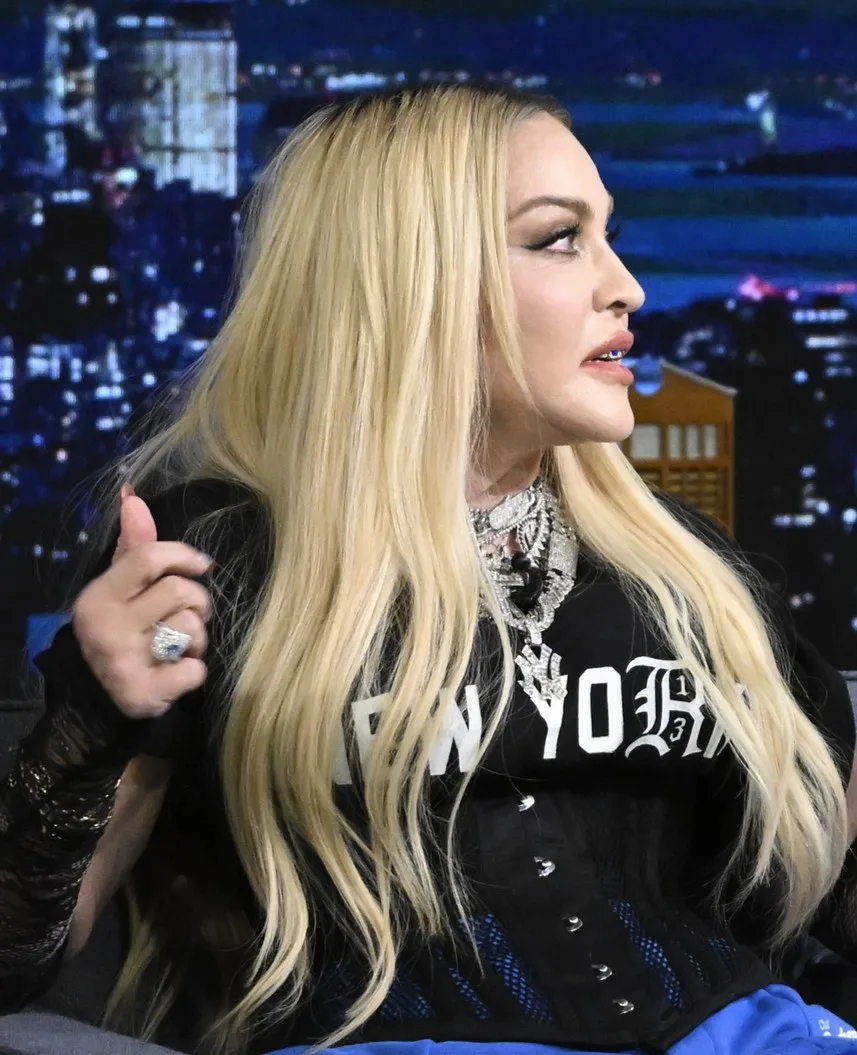 Madonna appears in profile during a television appearance with long blond hair, a black "New York" top, layered necklaces and a large statement ring.