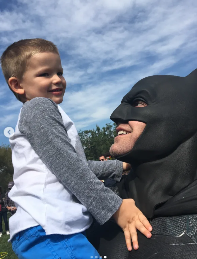 Ben Affleck, dressed in his Batman costume, shares a playful moment with his young son during an outdoor gathering. Fans were touched to see the throwback image included among Jennifer Garner's Father's Day memories, with many praising the former couple's enduring co-parenting relationship.